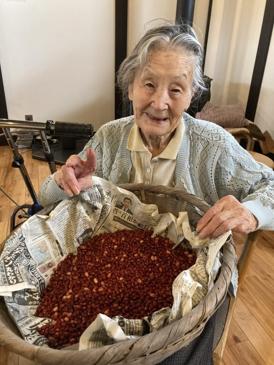 An elderly woman smiling behind a large basket of red azuki beans she has harvested, her walking frame just behind her