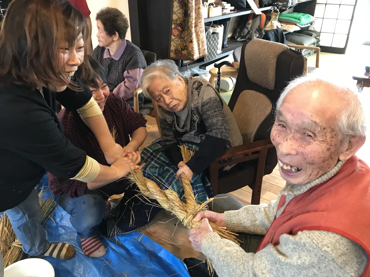 An elderly man laughing as he twists rice straw into rope, showing two younger caregivers how it is done