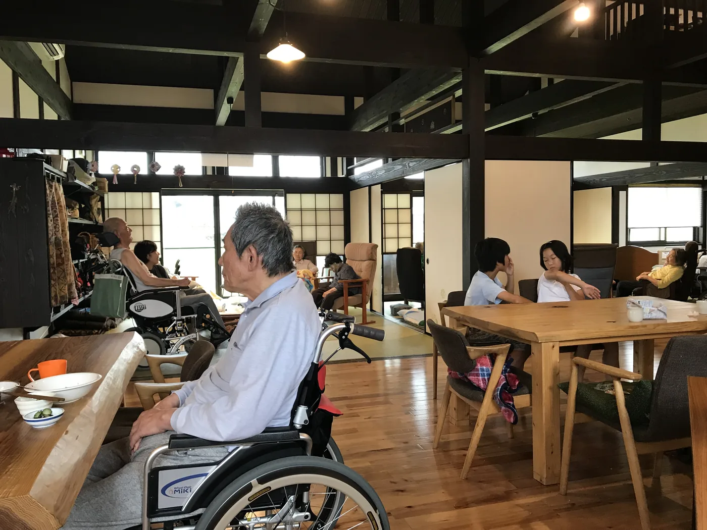 Inside the farmhouse: dark wooden beams, paper screens and a wide wooden floor, residents at tables and two people in wheelchairs
