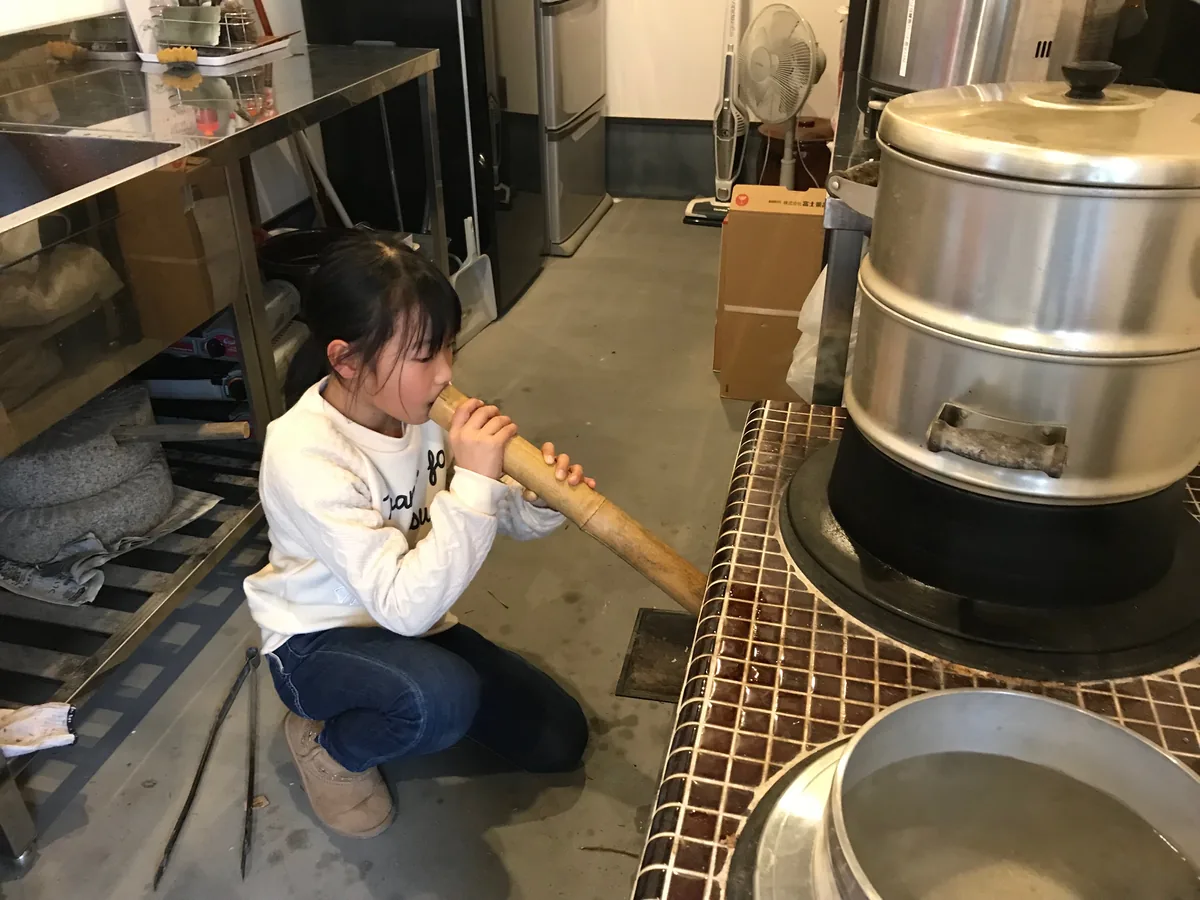 A child kneeling on the kitchen floor, blowing down a bamboo tube to feed the fire under a large steaming rice pot on a wood-fired okudo-san stove