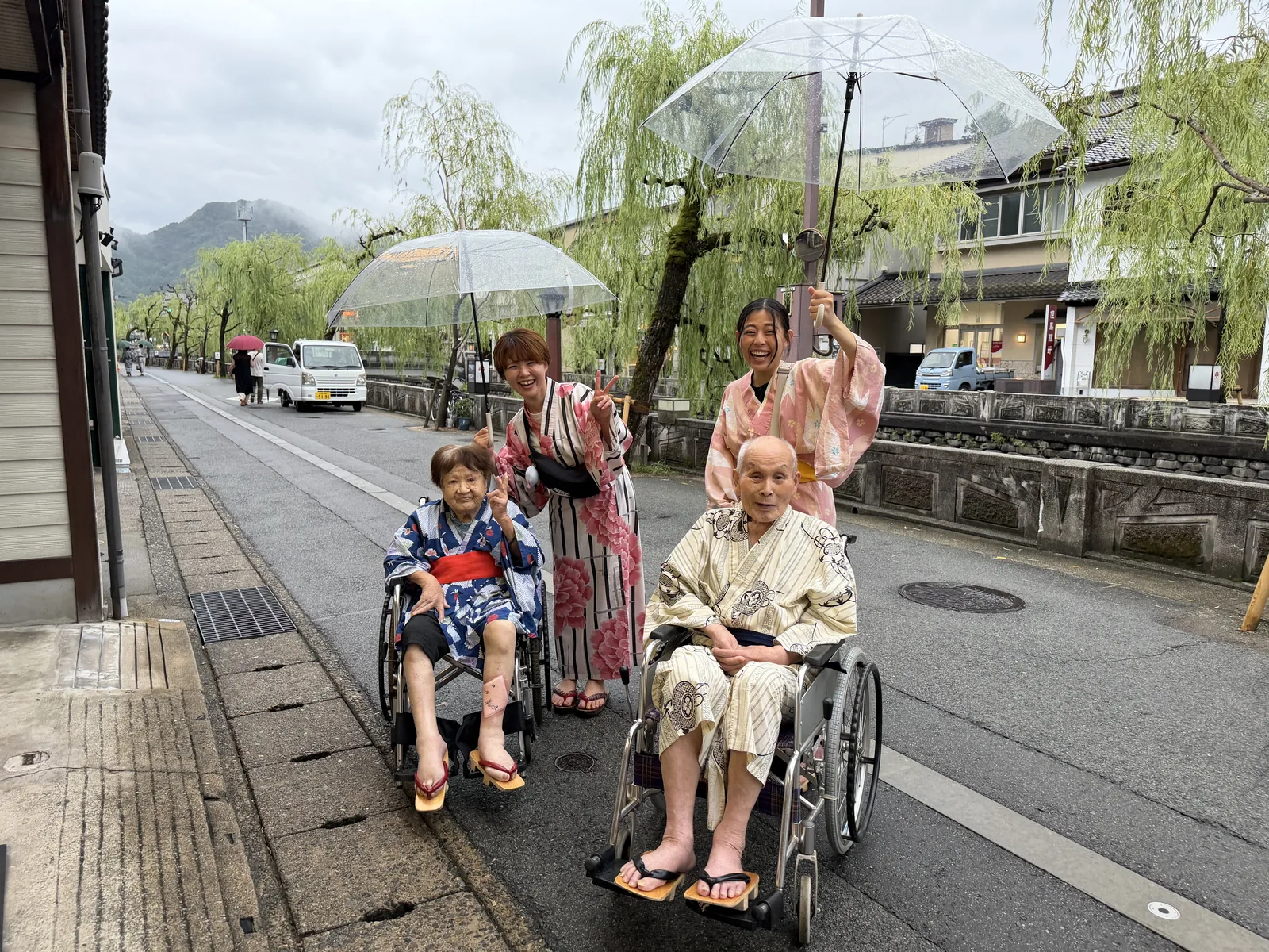 Two elderly people in wheelchairs wearing summer yukata, smiling under clear umbrellas on a willow-lined street, with two caregivers beside them